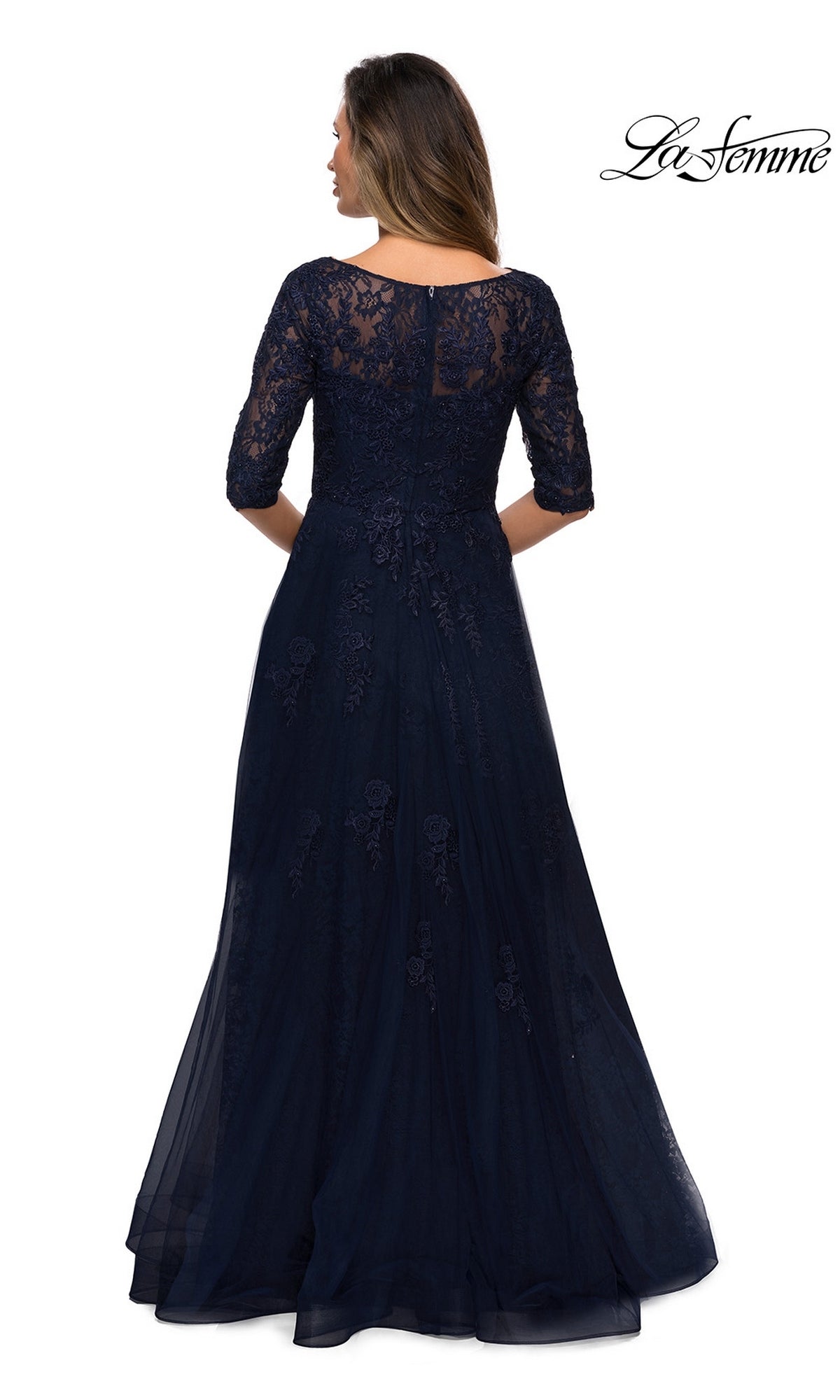 A woman in the Long Formal Dress: La Femme 28036 with navy lace, elbow sleeves, and a tulle A-line skirt stands facing away from the camera.